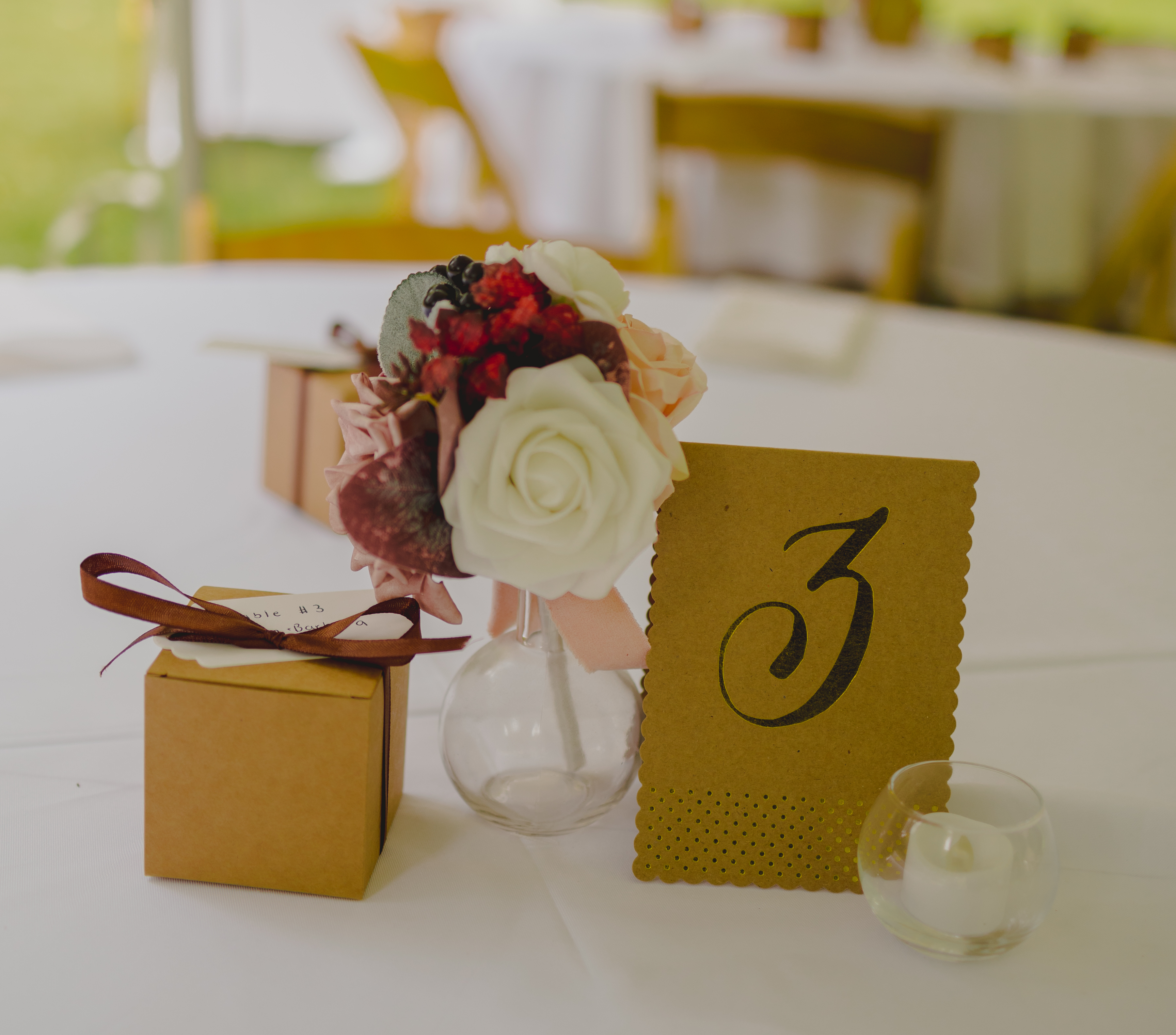 Simple decoration for a guest table at a wedding. The table has a vase of flowers, a table number, a gift box, and a small candle.